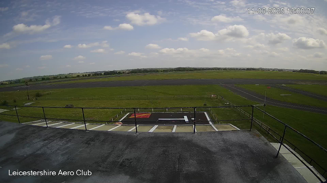 A wide view of an airfield under a partly cloudy sky, with a designated helicopter landing area marked with a large white "H" on a dark surface in the foreground. The airfield extends into green grassy fields and runways in the background. A timestamp in the upper right corner shows the date as June 11, 2025, at 12:00:37. The text "Leicestershire Aero Club" is visible in the lower left corner.