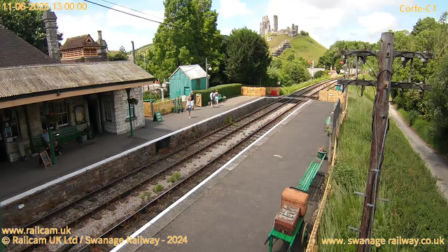 A small railway station platform with a stone building on the left side, featuring a tiled roof and a chimney. Several people are standing on the platform, some with luggage. A small green shed is behind them, and there is a green bench near the shed. The platform is bordered by a yellow fence at the far end and has a green bench on the right side. The railway tracks run between the platform and a grassy area with a narrow footpath on the right. Power lines with wooden utility poles run along the right side of the image. In the background, there is a hill with the ruins of a castle or old fortress on top, surrounded by trees. The sky is mostly clear with a few clouds. The timestamp on the image indicates the date as June 1, 2025, and the time as 1:00 PM.