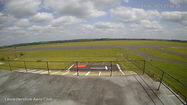 An outdoor view from a webcam at Leicesterhire Aero Club showing a clear sky with scattered clouds, an empty runway stretching across the distance, and a grassy area with some markings and fencing in the foreground. The timestamp indicates it was taken on June 11, 2025, at 1:00 PM.