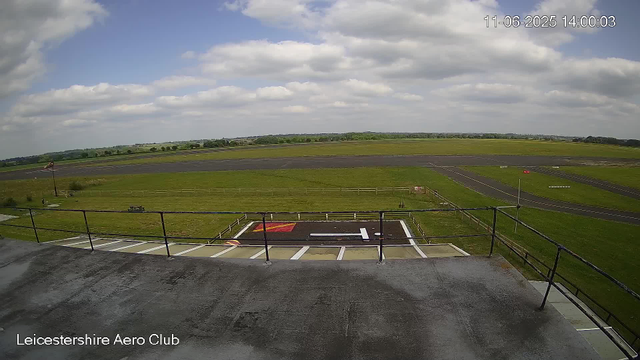 A view from a webcam at Leicestershire Aero Club shows an outdoor area with a grassy field, an empty tarmac runway, and a small section of parking or service area in the foreground. The sky is partly cloudy with a mix of white and gray clouds. The horizon features a distant landscape of green fields and trees. The image also displays a timestamp in the top right corner and the club's name in the bottom left corner.