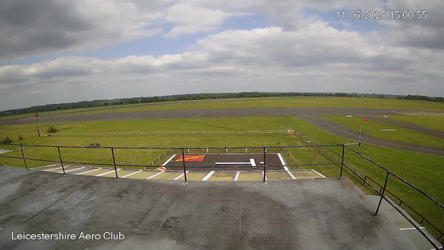 A view from a webcam at Leicestershire Aero Club shows an airfield with a small helicopter landing pad in the foreground. The pad has white and red markings, and a black railing surrounds it. Beyond the pad, there are grassy areas, a fence, and a runway extending into the distance under a partly cloudy sky. The timestamp in the top right corner indicates the date and time as 11th June 2025 at 15:00:55.