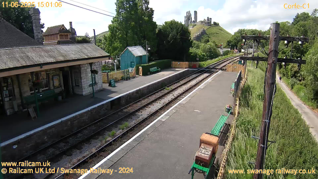 A railway station platform with a stone building on the left, featuring a roof with moss. There are green benches and flower pots along the platform edge. Tracks run diagonally from the foreground to the background. To the right, there are tall wooden poles supporting electrical wires. In the background, there are lush green trees, a small blue shed, a grassy hill with castle ruins on top, and a narrow pathway on the far right with a pedestrian walking. The sky is partly cloudy with patches of blue. The image includes a timestamp of June 11, 2025, at 15:01:06.