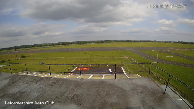 An outdoor view from a webcam at Leicestershire Aero Club showing a grassy airfield with a runway in the distance under a partly cloudy sky. The foreground includes a concrete balcony with a black railing and a marked helipad with a white "H" in the center and red markings. The horizon features a line of trees and open fields. The timestamp in the upper right corner reads "11-06-2025 16:00:57".