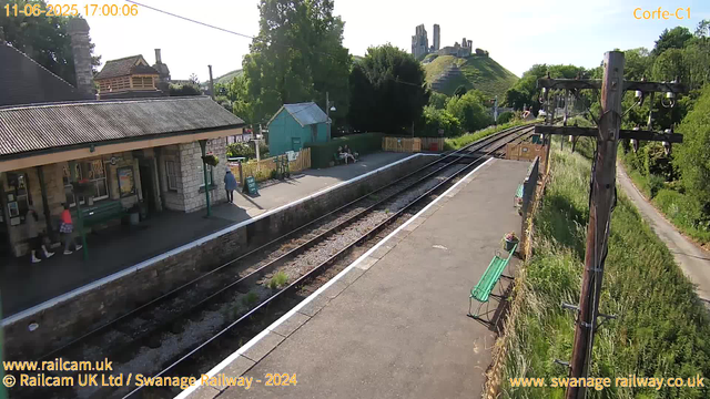 A small train station platform with a stone building on the left, two people sitting on benches, and one person standing near the entrance. There are two railway tracks running through the scene. To the right, there's a pathway with a green bench and potted plants, bordered by tall grass and bushes. Overhead, a wooden utility pole with power lines extends into the distance. In the background, a hill with castle ruins is visible, along with lush green trees under a clear sky. The timestamp indicates the date as June 11, 2025, at 17:00.