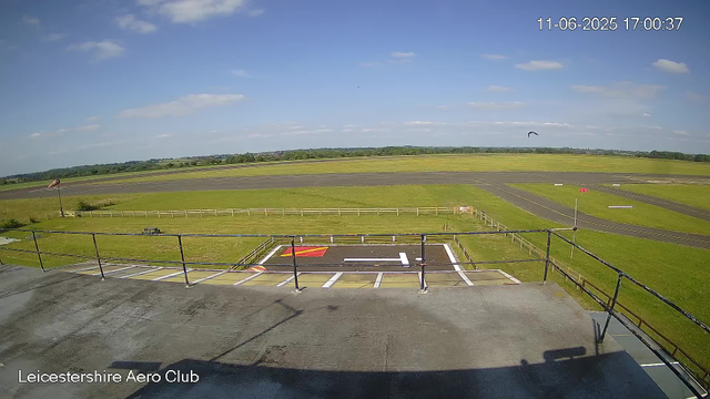 A clear view of an airfield with a grassy area, runway, and taxiways extending into the distance under a partly cloudy sky. A small fenced platform with a ramp and painted markings is visible in the foreground, with a windsock on a pole to the left. A bird is flying in the sky near the horizon. The top right corner shows the date and time as "11-06-2025 17:00:37". The bottom left corner displays the text "Leicestershire Aero Club."