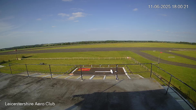 A view from a webcam at Leicesterhire Aero Club shows an empty tarmac area with a helicopter landing pad in the foreground, surrounded by a small grassy area with a wooden fence. Beyond the grassy area, there are multiple taxiways and runways extending into the distance under a blue sky with scattered clouds. The scene appears calm and sunny with no aircraft or people visible. The timestamp in the upper right corner reads "11-06-2025 18:00:21."