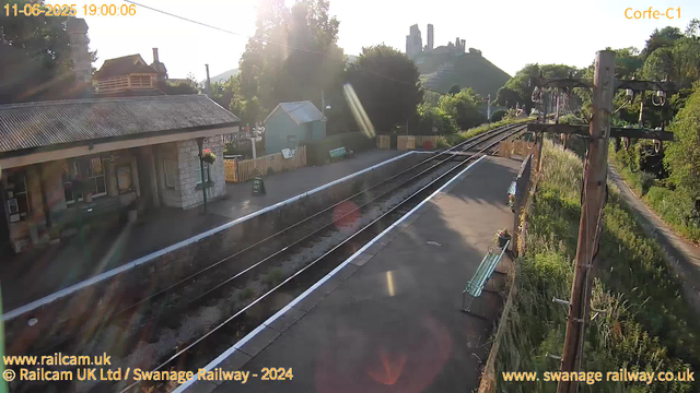 A small railway station platform bathed in early evening sunlight. The platform has a bench with flowers and a small building with a hanging flower basket. There are railway tracks running alongside the platform, with some overhead electric lines supported by wooden poles. In the background, there are green trees, a small house, and a hill with ruins or a castle. The sky is clear, and the overall scene appears calm and quiet. The timestamp indicates the date as June 11, 2025, at 7:00 PM.