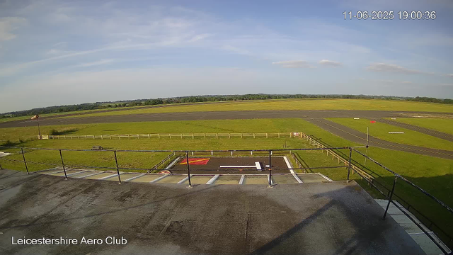 A view from a webcam showing the runway and surrounding grassy areas of Leicesterhire Aero Club. The sky is mostly clear with a few wispy clouds. The timestamp in the upper right corner indicates the date and time as June 11, 2025, at 19:00:36. There is a windsock on a pole to the left and some small markings on the tarmac indicating parking or taxi areas.