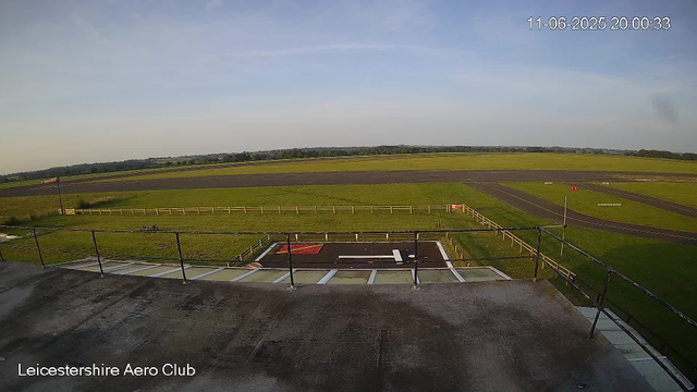 An empty helicopter landing pad with a white "H" in the center, surrounded by a black border. Fencing encloses the pad, and there is a small grassy area with a bench to the left. Beyond the fence, open green fields stretch toward the horizon under a clear blue sky. The timestamp in the top right corner reads "11-06-2025 20:00:33," and the bottom left corner has the text "Leicestershire Aero Club."