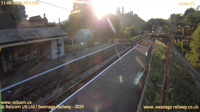 A small railway station platform with a building on the left, a bench, and a sign that says "WAY OUT." The railway tracks run parallel to the platform, and there is a wooden signal or utility pole on the right side with wires connected to it. The sun is low in the sky, creating a bright glare and lens flare effects, indicating it is either early morning or late afternoon. In the background, there are trees, a hill with ruins or a castle on top, and some residential houses with chimneys. The timestamp shows the date as June 11, 2025, at 8:01 PM.