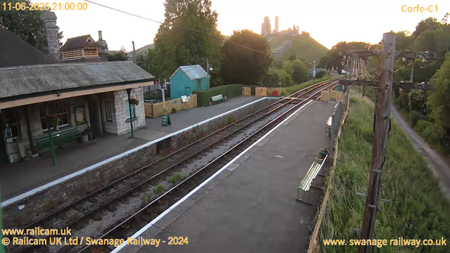 A small train station platform at sunset with train tracks running alongside. There is a stone building with a tiled roof and green benches under a covered area. A small green shed and a white fence are visible on the opposite side of the platform. Wooden pylons with overhead electrical wires run along the right side of the tracks. A winding footpath and trees are in the background, with a hill topped by historic structures silhouetted against the orange sky. The scene is quiet and empty of people.