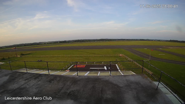 Looking out from a balcony over an airfield, the image shows a clear sky with some scattered clouds and contrails. In the foreground, there is a section of the balcony railing and a helipad marked with a large white "H" on a black background, with some red markings. The grassy airfield extends into the distance, with multiple runways and taxiways visible. A windsock is seen on the left side of the image. The timestamp in the top right corner indicates the date and time as June 11, 2025, at 21:00:41, and the bottom left corner has the label "Leicestershire Aero Club."