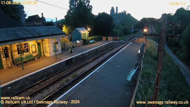 A small railway station platform at dusk with a green building on the left, benches, and a lamppost illuminating the area. Railway tracks run through the middle, extending into the distance. On the right side, there are utility poles with electrical wires, and a grassy area with a narrow path. In the background, there's a hill with ruins or old structures on top, partially obscured by trees. The sky is transitioning from light to dark, indicating evening time. The image is timestamped 22:00:29 on June 11, 2025.