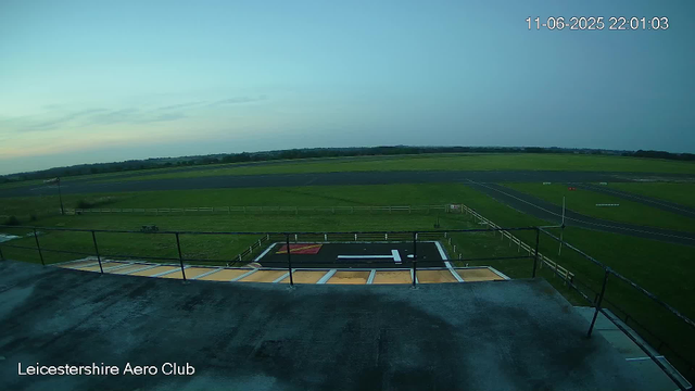 An empty airfield with a helicopter landing pad marked with a black and white "H" in the center. Surrounding the pad is a white fence, and the area appears to be part of the Leicester Aero Club. The scene shows a grassy field extending into the distance with a runway and taxiways, under a clear to partly cloudy sky during dusk or dawn. The timestamp on the image reads 11-06-2025 22:01:03.