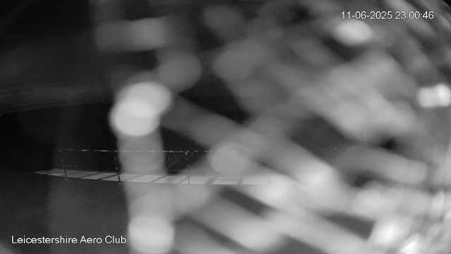 A black-and-white, blurry webcam image showing a dark outdoor area, possibly at night. There is a chain-link fence near the bottom of the image, and some out-of-focus lights in the background. The timestamp in the top right corner reads "11-06-2025 23:00:46," and the text in the bottom left corner indicates it is from the Leicesterhire Aero Club.