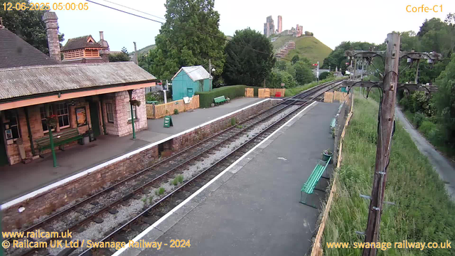 A small railway station platform with a stone building on the left, featuring green benches and hanging flower baskets. There are railway tracks running parallel to the platform, with a green bench and a small green shed in the background. A sign that reads "WAY OUT" is visible on the platform. On the right, there are wooden fences with potted plants and a row of electrical poles with wires stretching across the scene. In the background, a grassy hillside with a castle ruin perched on top can be seen. The scene appears to be early morning, as indicated by the timestamp.