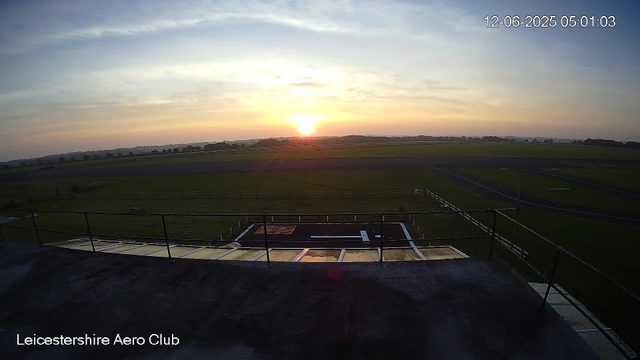 A sunrise over a flat, grassy landscape seen from a high vantage point at the Leicestershire Aero Club. The sky has soft, light clouds, and the sun is just above the horizon, casting a warm glow across the scene. In the foreground, there is a black rooftop with a safety railing and some equipment or panels visible. The open fields extend into the distance with a few trees dotting the horizon. The date and time display in the top right corner reads "12-06-2025 05:01:03".