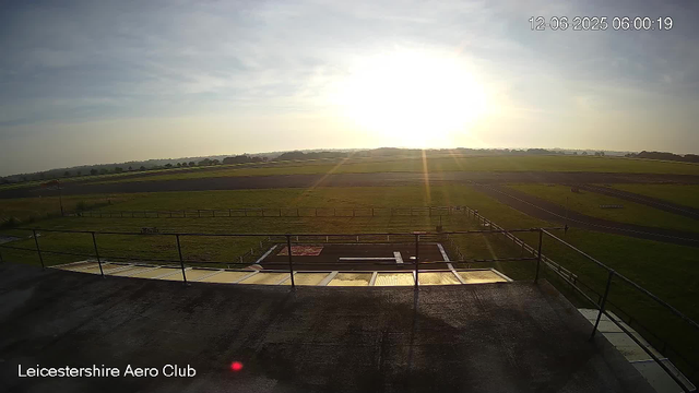 A wide-angle view of an open grassy field with a runway and fencing, captured by a webcam at sunrise or sunset. The sky is partly cloudy, and the sun is low on the horizon, casting a bright glow over the landscape. The image is marked with the timestamp "12-06-2025 06:00:19" in the top right corner and the label "Leicestershire Aero Club" in the bottom left corner.