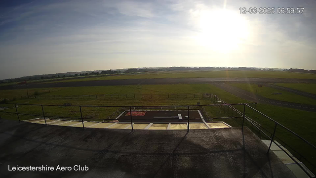 A wide-angle view from a webcam at Leicestershire Aero Club shows a grassy airfield with runways and taxiways stretching into the distance. The sky is partly cloudy with the sun shining brightly on the right side of the image. In the foreground, there's a platform or balcony with a black railing. The date and time in the upper right corner indicate December 6, 2025, at 6:59 AM.