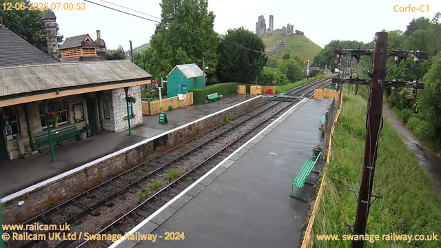 A small train station platform with a stone building on the left, featuring benches and hanging flower baskets. There are yellow safety barriers at the far end, along with green benches and a small teal shed. The railway tracks run through the center of the image, with greenery and trees on either side. In the background, a hill with a castle ruin is visible, and there are overhead electric wires and power poles lining the tracks. The sky is overcast, and the station area appears to be quiet.