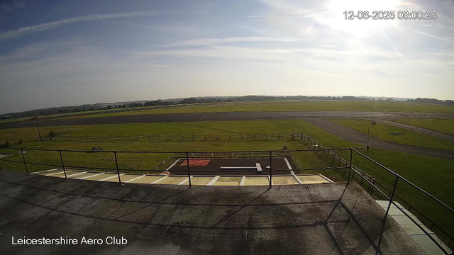 A daytime view from a webcam at Leicesterhire Aero Club shows a clear sky with some wispy clouds and bright sunlight. The scene includes a runway in the distance, a grassy area surrounding it, and a small helipad marked with a white "H" and red background in the foreground. There is a railing along the edge of what appears to be a balcony or platform. The image is timestamped "12-06-2025 08:00:25."
