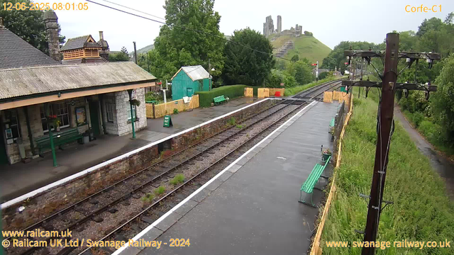 A small railway platform during daytime with an old stone station building on the left, featuring green benches and a shelter. The tracks run through the center, with a green shed in the background and a large hill with a ruined castle on top visible in the distance. There are several wooden barriers across the tracks, possibly indicating construction or maintenance. To the right, there are green benches along the platform and a narrow path with grassy following area beside a wooden fence and a railway line with overhead wires. The overall scene appears to be in a rural or semi-rural setting. The image has a timestamp of June 12, 2025, at 8:01 AM.