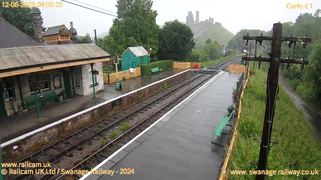 A small train station platform on a rainy day with empty green benches, a stone building with a sloped roof, and a small green shed surrounded by a yellow fence. There are trees and a castle on a hill in the background. A tall wooden utility pole with electrical wires is on the right side, and the train tracks run parallel to the platform. The sky is overcast, and the platform surface appears wet from the rain.