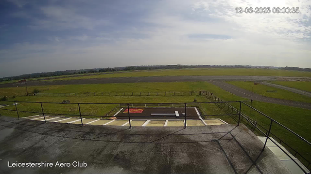A view from a webcam showing an outside scene at Leicestershire Aero Club. In the foreground, there is a platform with a safety railing and a small helipad with a white "H" marking. Beyond the platform, there is a grassy area with a flagpole on the left side. The scene extends to an open grassy field and a runway, with a clear sky overhead featuring some scattered clouds. The timestamp in the upper right corner reads "12-06-2025 09:00:35".