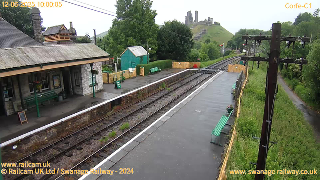 A small train station platform with benches and a green shed is visible. There are train tracks running alongside the platform, and a yellow safety barrier at the end of the platform. A large, grassy hill with a ruined castle on top is in the background. The weather appears overcast, and there is lush greenery surrounding the station. There are power lines and a utility pole on the right side of the image. The timestamp at the top left indicates the date and time as June 12, 2025, at 10:00:05.