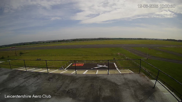 The image shows a wide view of an airfield from a webcam positioned on an elevated platform with a railing. In the foreground, there is a black helicopter landing pad with a white "H" symbol and a red square nearby, marked with white lines. The airfield extends into a grassy area and runways that stretch into the distance. The sky is partly cloudy with patches of blue, and the landscape is mostly flat with some distant trees and buildings. The time and date are displayed in the upper right corner: 12-06-2025 10:00:50. The bottom left corner has the label "Leicestershire Aero Club."