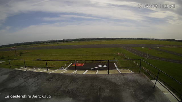 An outdoor view of an airfield with a grassy runway area, a helipad marked with an "X" and a red square, a fence, and a small vehicle parked near the fence. The sky is partly cloudy with patches of blue, and in the distance, there are flat fields and a tree-lined horizon. The timestamp in the top right corner shows "12-06-2025 11:00:20," and the bottom left corner displays "Leicestershire Aero Club."