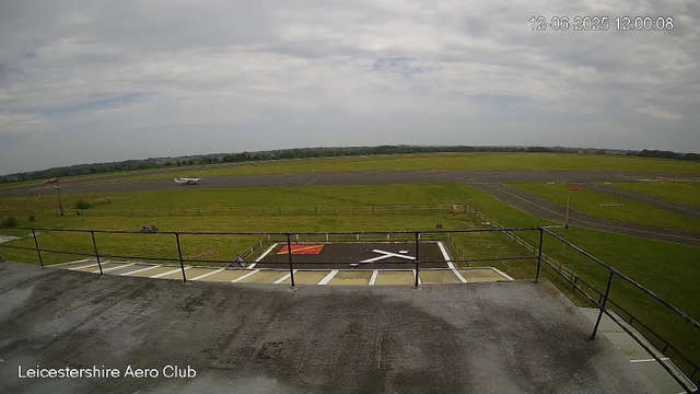 The image shows an airfield at the Leicestershire Aero Club on a cloudy day. In the foreground, there is a helicopter landing pad marked with a large black "H" inside a red square and crossed by white lines. A metal railing surrounds the area, and part of the rooftop from which the photo was taken is visible. Beyond the landing pad, there is a grassy field and a runway with a small airplane taxiing on it. The landscape extends into the distance with a flat horizon and a partly cloudy sky overhead. The timestamp indicates the date and time as December 6, 2025, at 12:00:08.