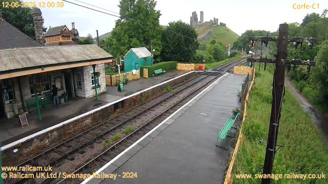 A small railway station platform with a stone building on the left, a person standing under the shelter, and empty green benches. There are railway tracks running through the center, with a wooden fence along the right side. In the background, a hill with ruins or a castle on top is visible, surrounded by green trees and shrubs. An old wooden utility pole stands to the right, with power lines stretching across the scene. The sky is overcast, and the timestamp indicates it is midday.