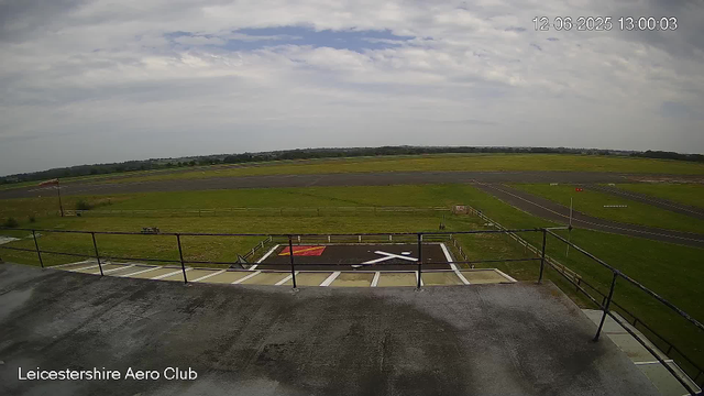 A view from a webcam at Leicestershire Aero Club showing a runway and grassy airfield area under a partly cloudy sky. The runway extends into the distance with some signage and markings visible. There's a small parking or helipad area in the foreground with a railing, and the landscape beyond consists of open fields and distant trees. The timestamp in the top right corner indicates the date as December 6, 2025, and the time as 13:00:03.