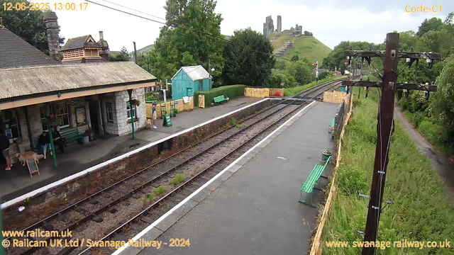 A small train station platform with a few people waiting, some sitting on green benches and one person standing with a dog. There is a stone building with a bench underneath the roof, and a small teal-colored shed nearby. Railroad tracks run parallel to the platform. In the background, a grassy hill with the ruins of a castle or large stone structure on top, partly obscured by trees. An old-fashioned electric pole stands on the right side, with wires hanging from it. There are some flower pots on the platform's edge, and a dirt path runs alongside the grassy area on the right side of the image. The timestamp indicates the photo was taken on June 12, 2025, at 1:00 PM.