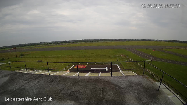 A view from a webcam shows the runway and surrounding grassy area of Leicesterhire Aero Club under a cloudy sky. The timestamp in the upper right corner indicates it was taken on June 12, 2025 at 13:59:53. The foreground includes a small helipad with a white "H" marking, enclosed by a black metal fence. The grassy landscape extends into the distance with a few small roads or pathways, and there is a windsock on a pole to the left side of the image. The overall scene appears calm with no aircraft or people visible.