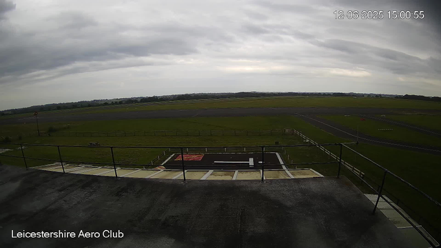 A view from a webcam showing the Leicester Aero Club's runway and surrounding grassy fields. The sky is overcast with gray clouds. The image is timestamped December 6, 2025, at 15:00:55. There is a fenced platform in the foreground overlooking the airfield.