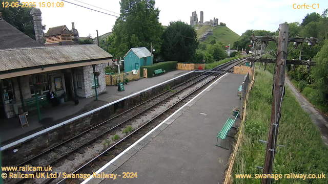 A small train station platform with a stone building on the left, where a person in dark clothing is sitting on a bench. There are green benches, signs, and potted plants. To the right, there are railway tracks running parallel to the platform, with grassy areas and trees surrounding the station. In the background, a hill with an ancient castle ruin on top is visible, along with some trees and greenery. There are electrical poles and wires along the platform, and the sky is partly cloudy.