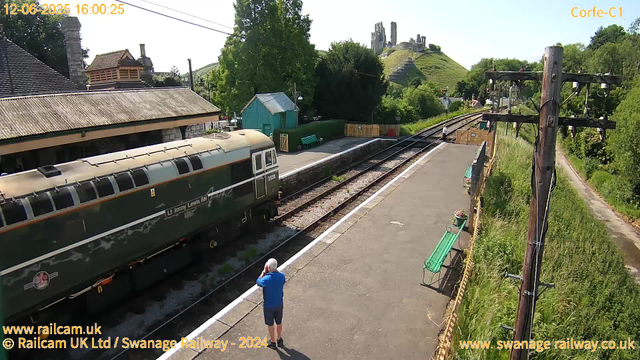 A vintage green train with a cream roof is stopped at a small railway station platform, with a man in a blue shirt taking a photo of it. There are benches with flower pots, a small green building, and tall utility poles beside the platform. In the background, a hillside with the ruins of an old castle or fortress is visible, and a tree with lush green foliage partially covers nearby houses. The sky is clear and blue, indicating a bright, sunny day.