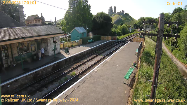 A small train station with a platform on the left side, where two people are standing near a building, and an empty bench nearby. There are railway tracks running through the middle of the image, curving into the distance. On the right, there is a wooden utility pole with wires, and tall grass and vegetation alongside the platform. In the background, a hill with the ruins of an old castle or structure is visible, surrounded by trees and greenery. The sky is clear and sunny. The image has timestamps and website credits at the top and bottom.