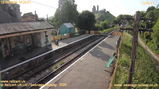 A small train station platform sheltered by a stone building with a pitched roof. There are two green benches on the platform, and a small teal-colored shed nearby. Railroad tracks run through the scene, with grassy areas on both sides. In the background, there is a hill with the ruins of an ancient castle atop it. Utility poles with wires run parallel to the tracks. The sky is mostly clear with bright sunlight. The date and time displayed at the top left corner indicate it is June 12, 2025, at 6:00 PM.
