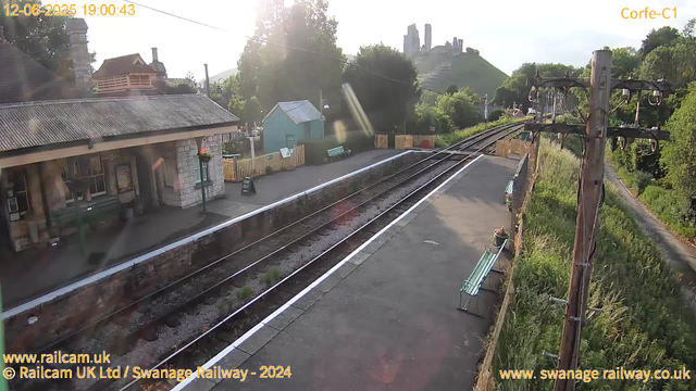 A quiet train station platform with no trains visible. To the left, there is a small stone building with a corrugated metal roof, and next to it is a flower basket hanging from a pole. In the background, a small turquoise building can be seen, with a fenced area and benches. On the right side, there are wooden utility poles with electrical wires, and a narrow path with grass and bushes runs alongside. In the distance, a hill with a historic castle or fortress ruins sits atop, partially immersed in sunlight. The overall scene appears peaceful with greenery and a clear sky. The date and time at the top left corner show "12-06-2025 19:00:43".