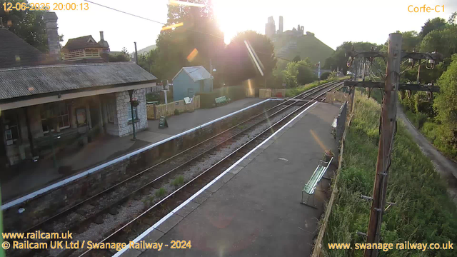 A small train station platform bathed in warm sunlight with the sun low in the sky. There are railway tracks running parallel to the platform, which appears to be empty. On the left, a building with a stone facade and a window, and a small shelter with a roof, are visible. There are benches along the platform, one with a flower pot. On the right, a tall wooden utility pole with multiple wires extending from it runs alongside a narrow grassy path bordered by lush green trees and bushes. In the background, a hill with a castle or large historic building is visible, illuminated by the setting sun. The date and time are displayed in the top left corner, and there are website and copyright notices at the bottom.