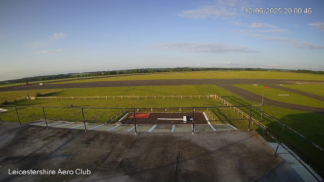 An outdoor view from a webcam at Leicesterhire Aero Club showing a clear sky with a few scattered clouds. In the foreground, there is a balcony or observation deck with a black railing. Below, there's a small tarmac area with markings and a section of grassy land behind a wooden fence. The landscape includes open grassy fields and a runway extending toward the horizon. The image is timestamped 12-06-2025 at 20:00:46.