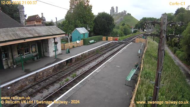 A small train station platform with a building on the left, featuring stone walls, a tiled roof, green benches, and a green lamppost. There are several empty green benches along the platform, with a sign indicating "Way Out." To the right, a set of railway tracks runs parallel, with a wooden utility pole and power lines overhead. In the background, a lush green hillside is visible, topped by a castle ruin on a hill. A paved path runs along the platform's right side, separated by a wooden fence with small flower planters. The sky is overcast. The image is timestamped "12-06-2025 21:00:37" at the top left.