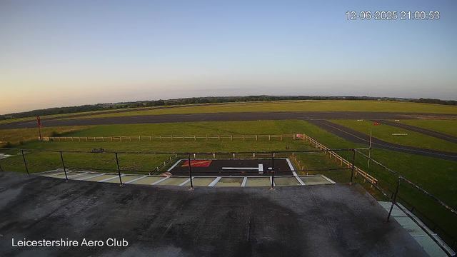 A view from a webcam at Leicesterhire Aero Club shows the edge of an aircraft runway in the distance, with a grassy area and a small paved area with markings in the foreground. The scene is set during early evening with a clear sky, and the timestamp indicates it is June 12, 2025, at 9:00 PM.