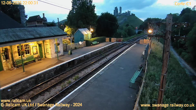 A small train station platform in a rural area during dusk. The platform has a green bench, a small green building, and several hanging flower baskets. There are railroad tracks running alongside the platform with a row of wooden fencing and a large electric pole with electrical wires. In the background, there is a hillside with a castle ruin on top, and the surrounding landscape is lush and green. The sky is mostly cloudy with some fading daylight, indicating early evening. There are a few street lamps lighting the platform.