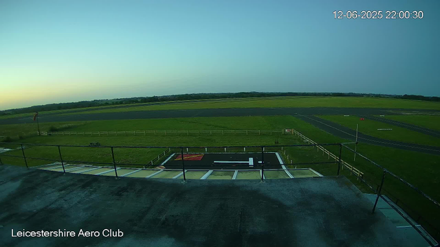 This image shows an empty aircraft runway with a small landing pad or helipad in the foreground, marked with a large white "H" inside a black square. Surrounding the runway are grassy fields extending into the distance. In the upper right corner, there is a timestamp reading "12-06-2025 22:00:30". The bottom left corner has the text "Leicestershire Aero Club". The scene is captured during dusk or early evening, with a clear sky. A windsock is visible to the left, and a white fence surrounds the landing area.