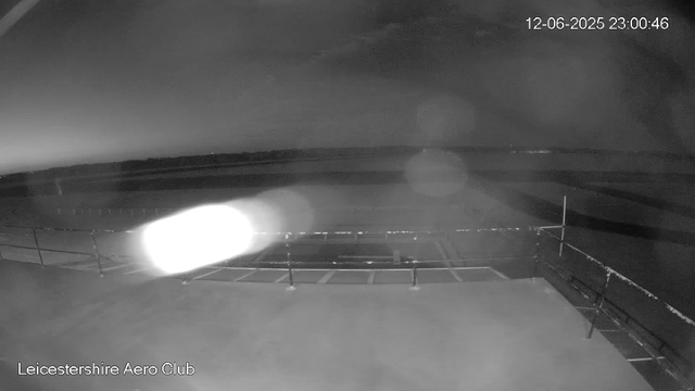 A black-and-white nighttime webcam image shows an empty tarmac or parking area at Leicesterhire Aero Club. The sky is mostly dark with some faint cloud formations. Bright light glare appears near the bottom left corner, possibly from the camera flash or headlight. There are a few parked cars and a fence at the edge of the area. The time and date displayed in the top right corner are December 6, 2025, at 11:00 PM and 46 seconds.