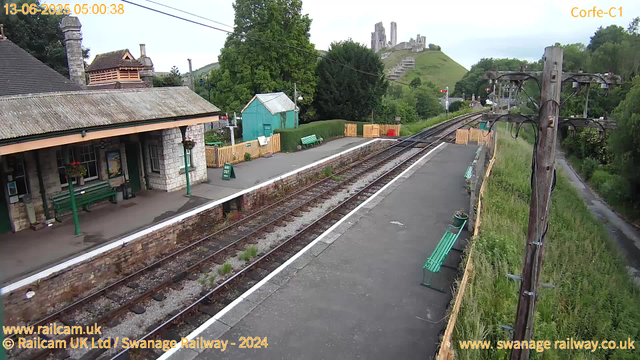 A vintage railway station platform with stone and brick buildings on the left, featuring green benches, planters, and a "Way Out" sign. Tracks run down the middle, bordered by a low brick wall. Several green benches are placed along the platform, and a small teal shed is visible behind a neatly trimmed hedge. Overhead, a few wires stretch across the scene. In the background, a lush green hill rises with an ancient castle ruin atop, and trees surround the area. The image appears to be captured early in the morning, as indicated by the timestamp.
